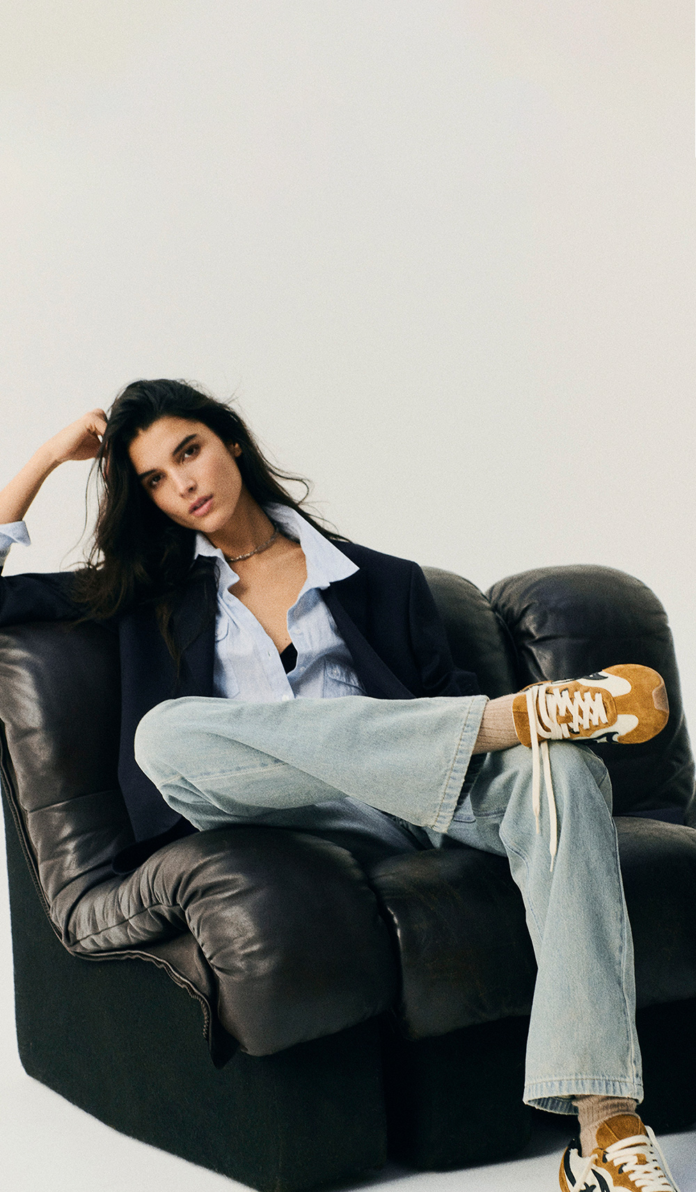 A model sitting on a black sofa wearing a denim outfit, a black blazer and Ba&sh sneakers.